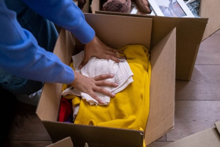 person packing folded clothes into a cardboard box while decluttering a room