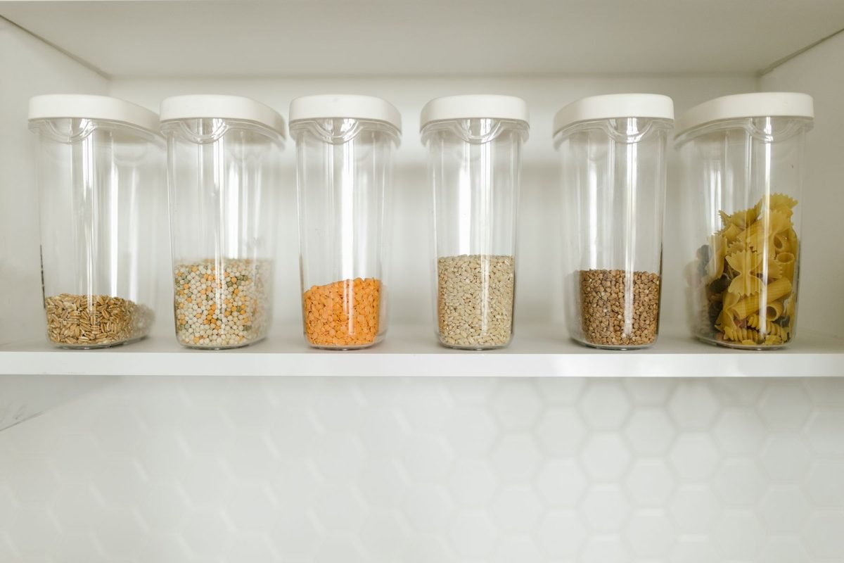 row of clear plastic containers holding grains, pasta and seeds on a white shelf